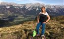 Eine Frau steht auf einem Berg mit einer atemberaubenden Aussicht auf die Alpen. Im Hintergrund sind schneebedeckte Gipfel und grüne Täler zu sehen.
