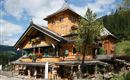 Ein traditionelles, holzverkleidetes Alpenhaus mit vielen bunten Blumen und einer malerischen Umgebung. Der Himmel ist blau mit einigen Wolken und grünem Wald im Hintergrund.