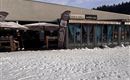 Een après-ski restaurant met een terras in de sneeuw. Er zijn parasols en de omgeving is winters en bergachtig.