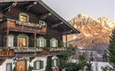 A traditional wooden house in the snow with balconies and flowers. Snow-covered mountains can be seen in the background.
