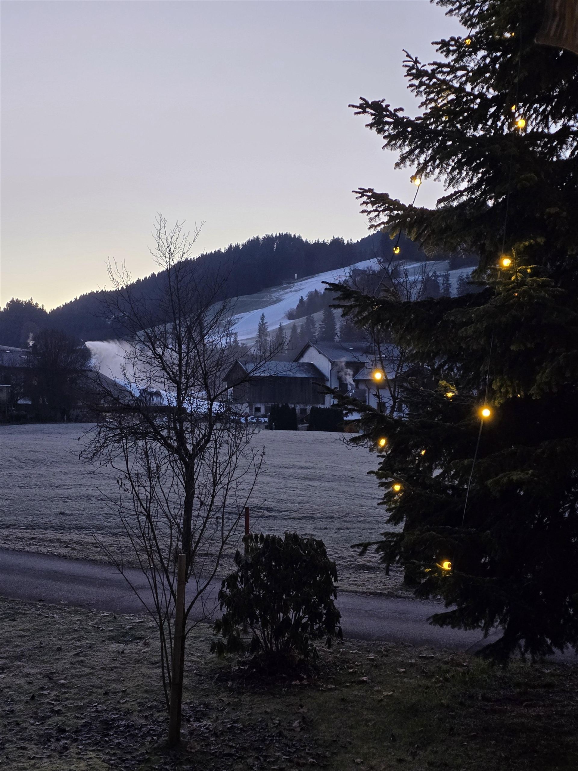 Ein winterlicher Morgen mit schneebedeckten Hügeln und einem Dorf im Hintergrund. Ein Tannenbaum mit Lichtern steht im Vordergrund.