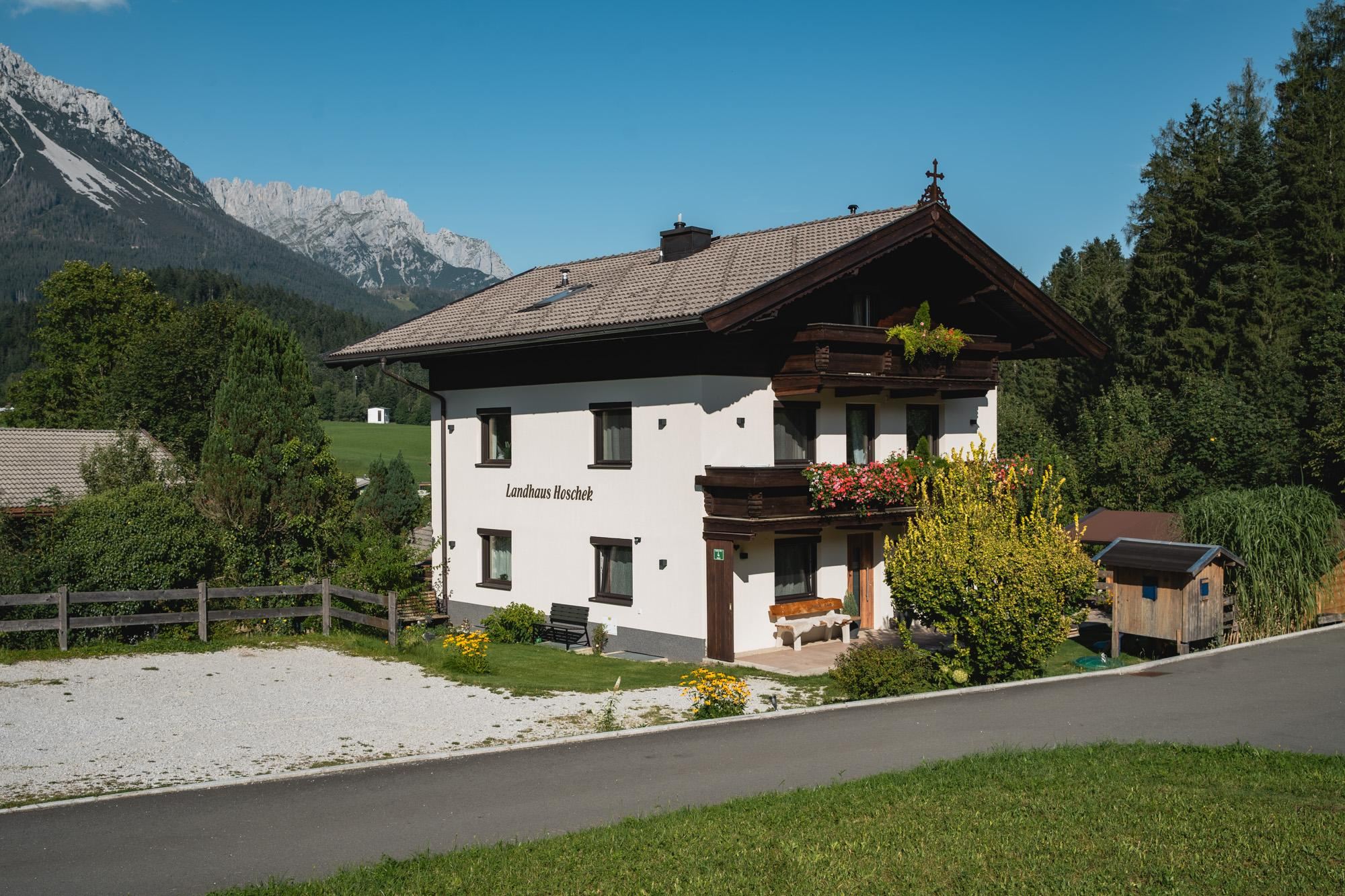 Ein hübsches, zweigeschossiges Haus mit traditioneller Architektur in einer grünen Landschaft. Im Hintergrund sind Berge und ein klarer blauer Himmel zu sehen.