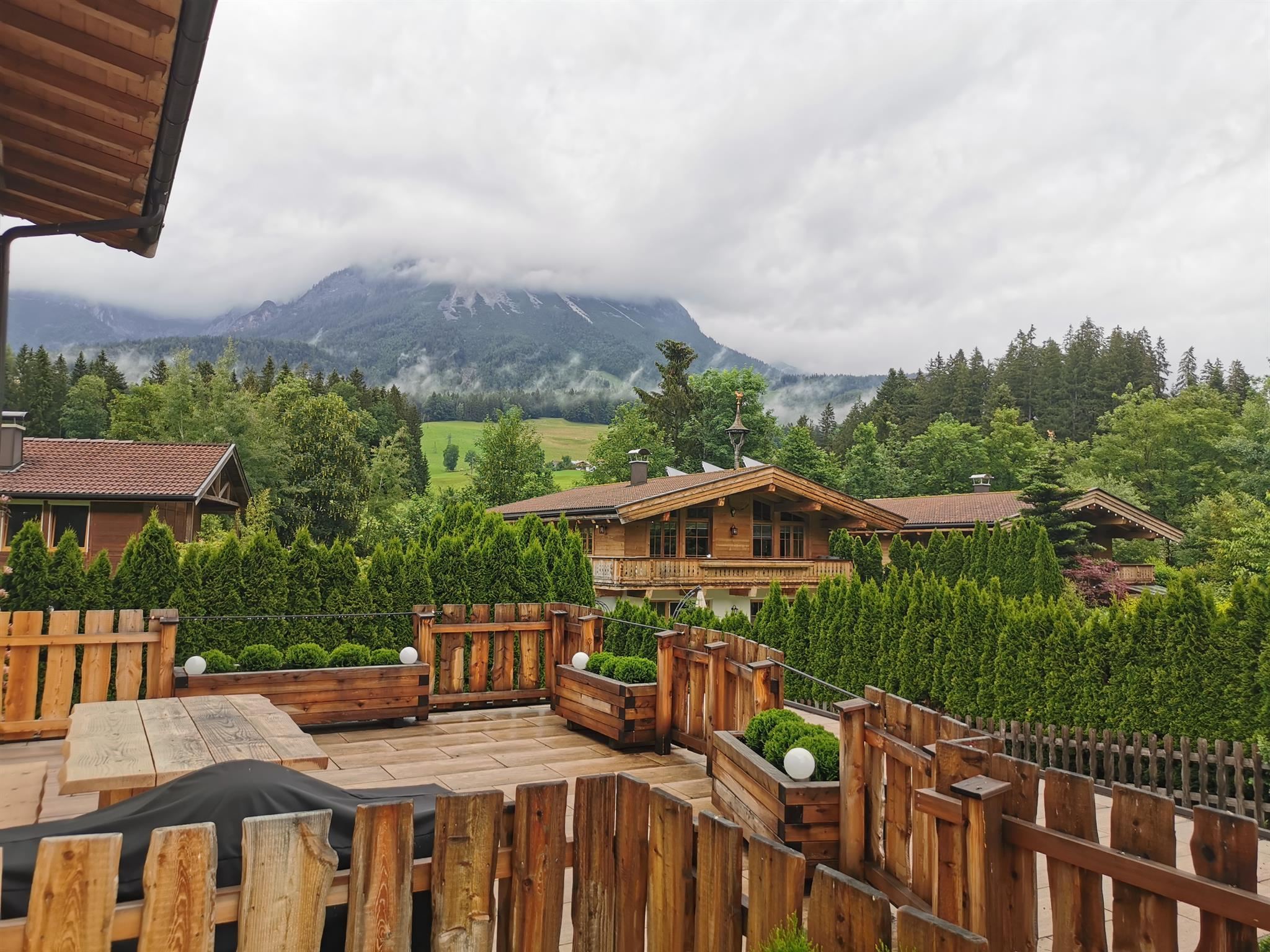 Eine ruhige alpine Landschaft mit Holzterrassen und typisch bayerischen Chalets. Im Hintergrund sind bewaldete Berge und leicht bewölkter Himmel zu sehen.