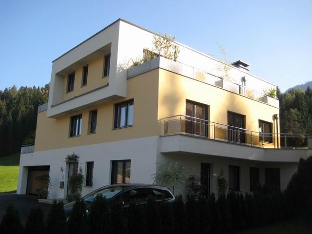Ein modernes, mehrstöckiges Haus mit gelber Fassade und einem Balkon. Es liegt in einer grünen Umgebung mit Bergen im Hintergrund.