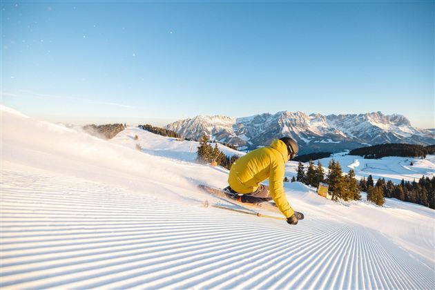 Ein Skifahrer trägt eine gelbe Jacke und fährt die perfekt präparierte Piste hinunter. Im Hintergrund sind schneebedeckte Berge und ein klarer blauer Himmel zu sehen.