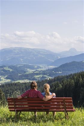 A couple is sitting on a bench, looking at a picturesque mountain landscape. The sky is clear and nature is lush green.