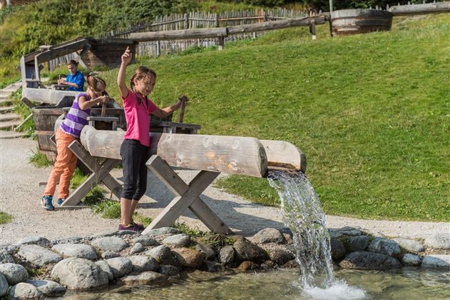 Children are playing by a stream outdoors. There are wooden structures and a green meadow in the background.