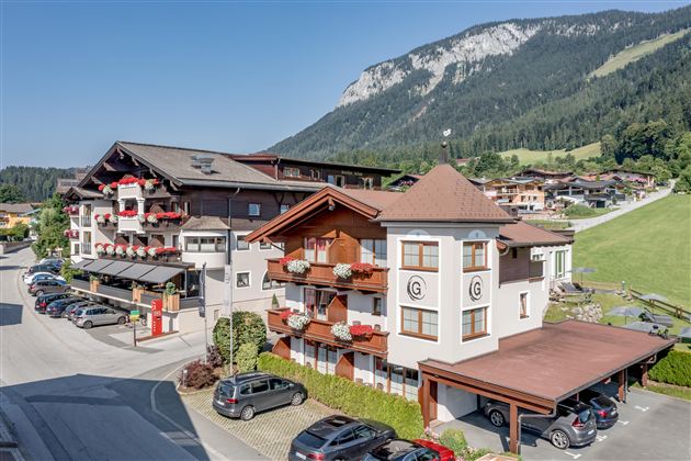 A picturesque Alpine landscape with several buildings in the foreground. The surroundings are green and mountainous, ideal for vacationers.