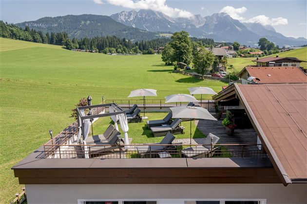 A rooftop terrace with sun loungers and umbrellas offers a beautiful view of green meadows and the mountains in the background. The environment is quiet and inviting.