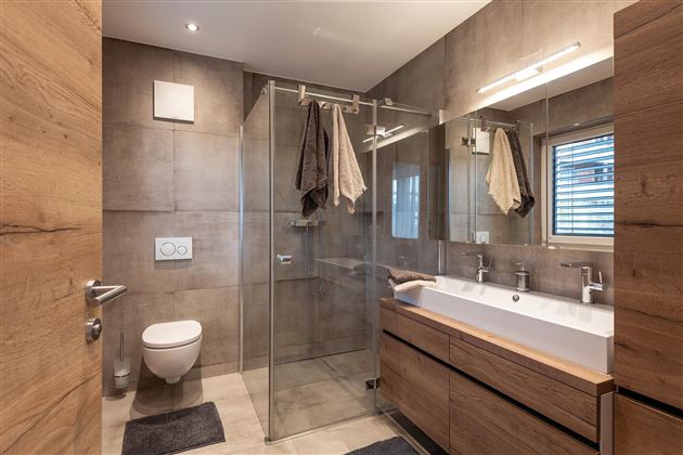 A modern bathroom with a glass shower cabin and wooden shelves. The walls are in neutral colors, and there are large windows for natural light.