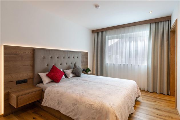 A modern bedroom with a large double bed and an elegant headboard. The room is bright and cheerful, with wooden flooring and curtains made of light fabric.