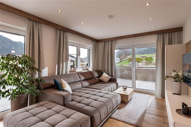 A modern living room with a comfortable gray couch and large windows. The room is bright and welcoming, with a view of a green landscape.