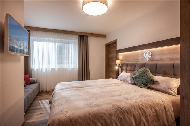 A modern bedroom with a large bed and a bright window front. The walls are in warm tones and there is a cozy seating area.