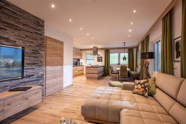A modern living room with a comfortable sofa and wood-paneled walls. The open kitchen is bright and inviting, with large windows and plenty of daylight.