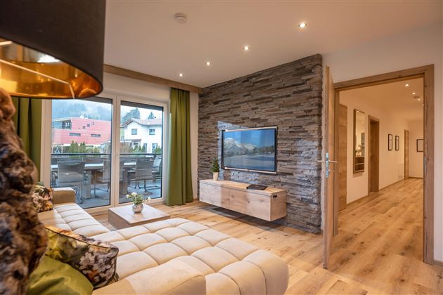 A modern living room with a cozy sofa and a television. Large windows offer a view of a balcony and plenty of daylight.