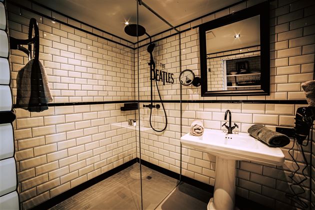 A modern bathroom with white tiles and a glass shower. On the wall, it says "THE BEATLES," and there are towels as well as an elegant washbasin.