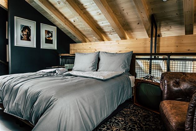 Ein stilvolles Schlafzimmer mit einem großen Bett und einem modernen Braun-Lederstuhl. Die Holzbalkendecke und die Bilder an der Wand verleihen dem Raum Wärme und Gemütlichkeit.