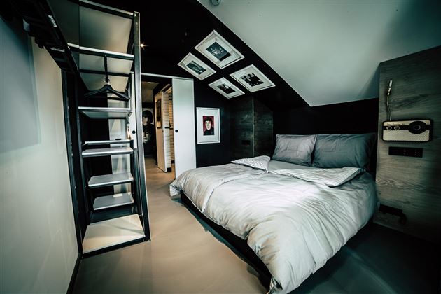 A modern bedroom with a large bed and a minimalist design. Photos hang on the wall, and there is an open wardrobe.