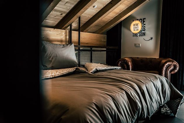 Ein gemütliches Schlafzimmer mit Holzbalken und einem großen Bett. An der Wand hängt eine Lampe mit einem inspirierenden Spruch.