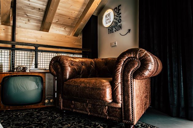 Ein gemütlicher, brauner Ledersessel steht in einem modernen Raum. Im Hintergrund befindet sich ein retro Fernseher und eine schöne Holzdecke.