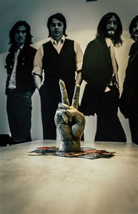Eine Hand mit dem Peace-Zeichen steht auf einem Tisch. Im Hintergrund sind Silhouetten von vier Männern zu sehen.