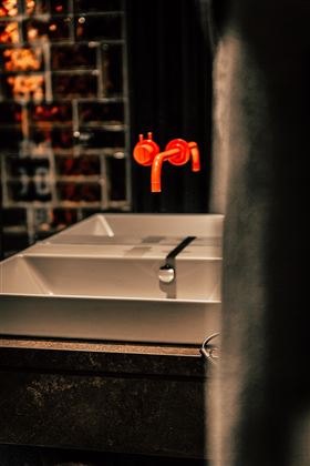 Ein modernes Waschbecken mit einem klaren, weißen Design und auffälligen, roten Wasserhähnen. Im Hintergrund sind stilvolle, dunkle Fliesen sichtbar.