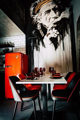 Ein stilvolles Esszimmer mit einem orangefarbenen Kühlschrank und einem auffälligen Wandgemälde. Der Tisch ist elegant gedeckt mit schwarzen und roten Stühlen.