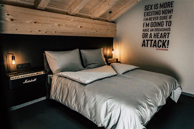 Ein modernes Schlafzimmer mit einem großen Bett und Holzbalken an der Decke. An der Wand befindet sich ein humorvolles Zitat und ein Verstärker in der Ecke.