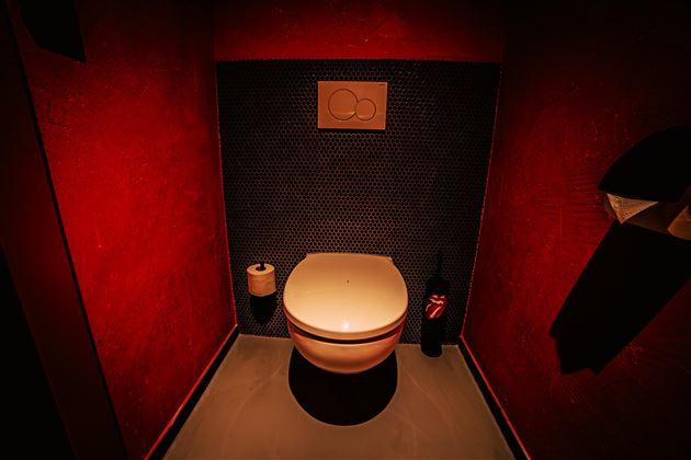 Ein modernes Badezimmer mit einem weißen Toilettenbecken vor einer rot gestrichenen Wand. Die Beleuchtung ist gedämpft und schafft eine intime Atmosphäre.