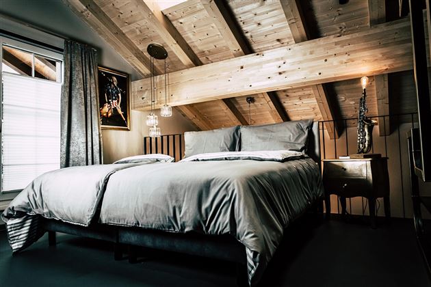 A stylish bedroom with a large double bed and wooden ceilings. The room has large windows and a cozy atmosphere.
