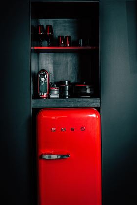 Ein roter Kühlschrank von SMEG steht in einer dunklen Küchenumgebung. Daneben sind einige Küchengeräte und Geschirr auf einem Regal zu sehen.