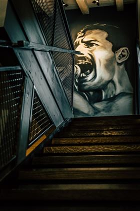 A staircase with dark wooden steps leads up. On the wall is a large, vivid mural of a screaming man.