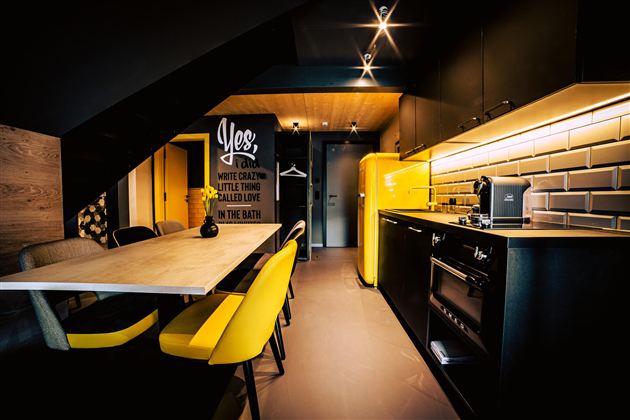 A modern kitchen with black cabinets and a yellow refrigerator. The dining area is equipped with a table and yellow chairs.