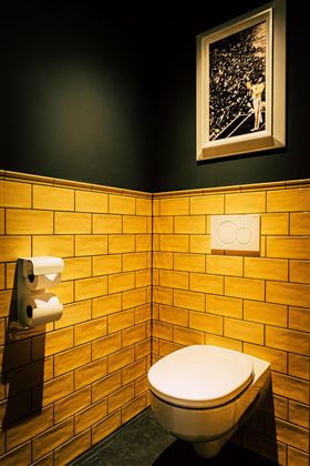 Ein modernes Badezimmer mit gelben Kacheln und schwarzer Wandfarbe. An der Wand hängt ein Bild, und es gibt eine Toilette mit Papierhaltern.