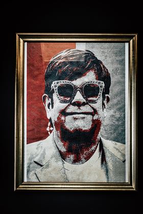 Ein künstlerisches Porträt einer Person mit Sonnenbrille und einem fröhlichen Ausdruck. Der Hintergrund ist in Rottönen gestaltet.