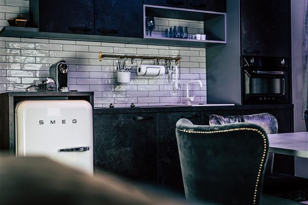 A modern kitchen with a stylish SMEG refrigerator and elegant black furniture. The walls are clad with white tiles, creating a minimalist look.
