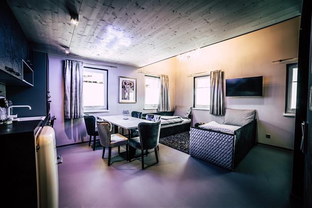 Ein modernes Wohnzimmer mit rechteckigem Tisch und bequemen Stühlen. Große Fenster lassen viel Licht herein und es gibt einen Fernseher an der Wand.