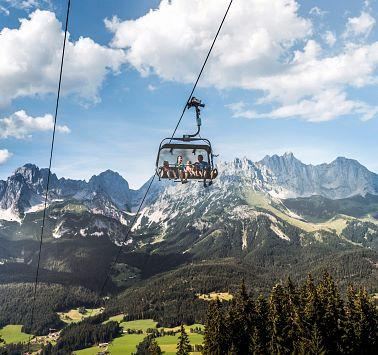 A cable car glides over green meadows and majestic mountains. The sky is clear and the landscape is stunning.
