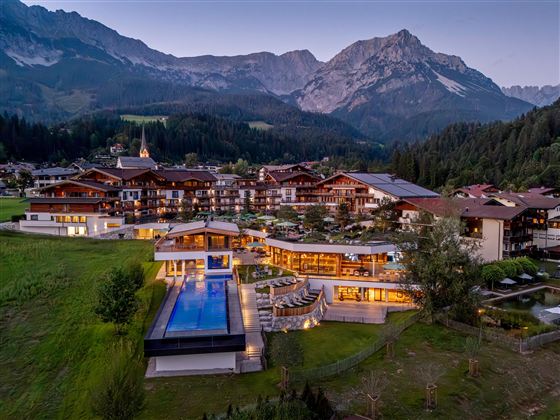 A picturesque hotel in the mountains with modern architecture and a beautiful pool. The surroundings consist of green meadows and majestic mountains.