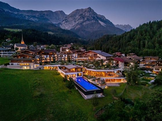 A picturesque landscape with a hotel complex, surrounded by mountains and forests. The outdoor area features a pool that is illuminated by lights.
