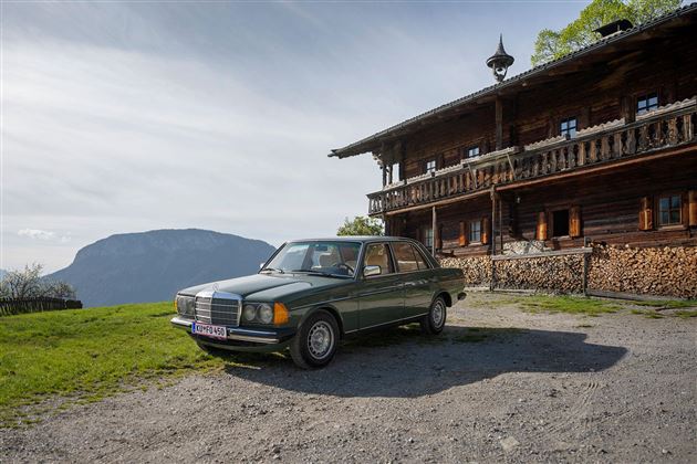 Ein klassisches Auto steht vor einem traditionellen Holzhaus in einer landschaftlichen Umgebung. Im Hintergrund sind Berge und ein klarer Himmel zu sehen.