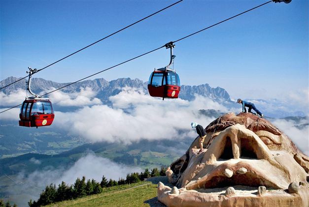 An impressive mountain landscape with gondolas floating above the clouds. In the foreground stands a large sculpture of a face.
