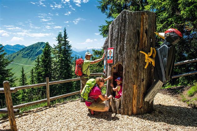A wooden sculpture in the shape of a tree with a colorful bird and an entrance for playing. Two hikers, an adult and a child, are happily interacting with the sculpture in the woods.