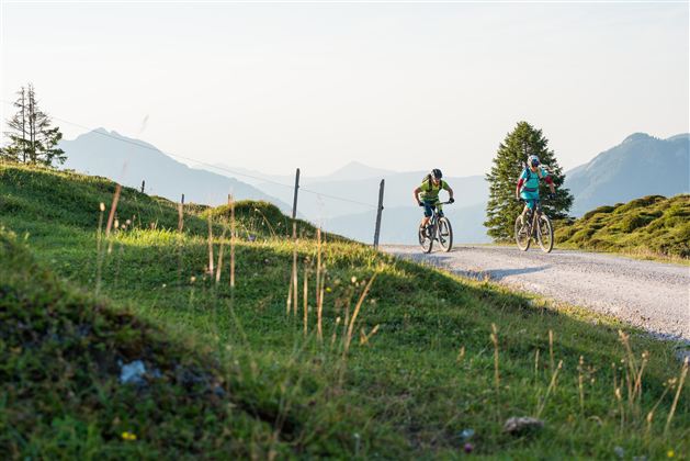 Two cyclists are riding on a gravel path through a green landscape. In the background, gentle hills and mountains can be seen.