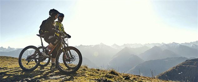 Two cyclists are standing on a high trail, admiring the mountain landscape. The sun is shining over the peaks, providing a breathtaking view.