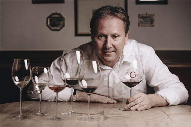 A man is sitting at a table with several wine glasses in front of him. He is holding a glass of wine in his hand and looking at the camera.