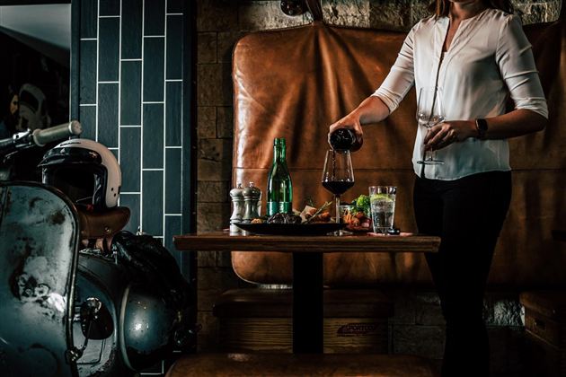 A person is pouring wine into a glass at a table with various drinks and snacks. In the background, there is a stylish decor with a leather bench and a motorcycle.