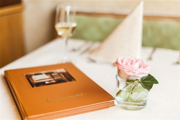 An elegant table with a menu and a rose in a glass vase. In the background, there is a glass of wine and a folded napkin on the table.