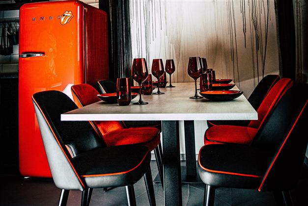 A stylish dining area with a white table, black and red chairs, and a striking red refrigerator in the background. Glassware and plates are beautifully set, adding a vibrant atmosphere to the space.