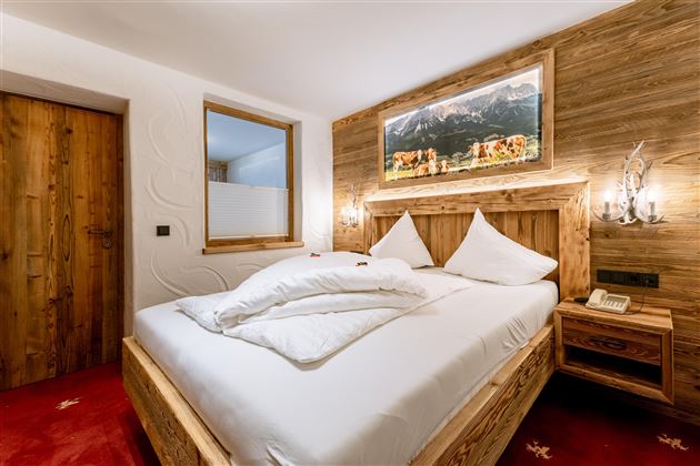 A cozy bedroom with a wooden bed and white pillows. A landscape picture hangs on the wall, and the floor is covered with a red carpet.
